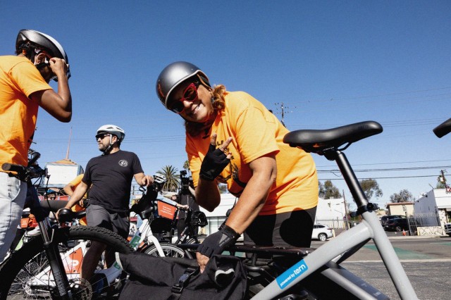 Woman of color bending over her bicycle giving a peace sign. She wears a bike helmet, sunglasses, and bike gloves. Two other bikers stand to her right getting their bikes ready.