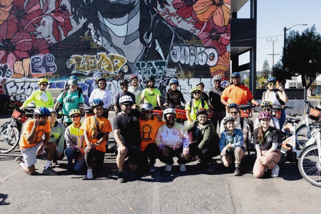 Two dozen adult bicyclists, some standing, some kneeling, posing with their bikes in a parking lot in front of a mural of Bob Marley surrounded by orange flowers.