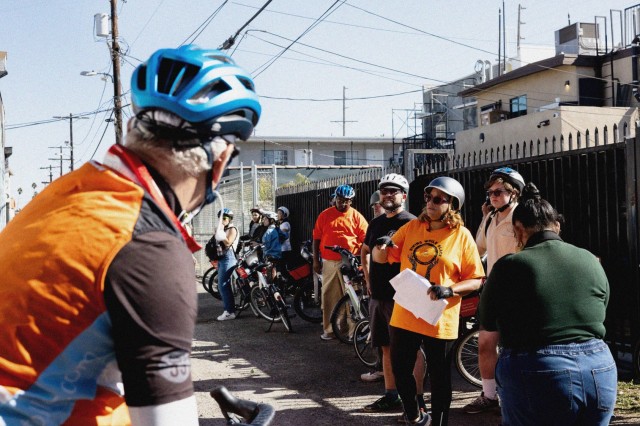 A group of a dozen BIPOC bike riders parked on the sidewalk in front of a black metal fence, circled around their group leader giving instructions.