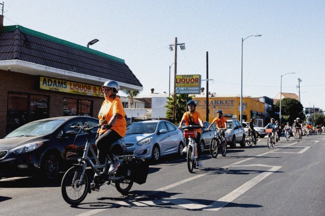 A line of bike riders riding in a bike path past parked cars and local stores.