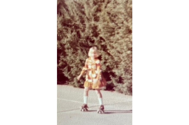 Young blond girl in an orange dress with white knee-high socks roller skating in a driveway in front of a row of tall bushes.