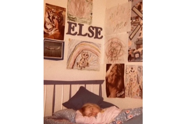 A young girl, blond, wearing pink pajamas, asleep in bed, with childhood drawings and posters on the wall around her bed. The name ELSE is spelled out on the wall.