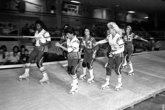Black and white photograph of eight women roller skating around a banked, wooden track. One team of four women wear a light-colored uniform, while the other team wears a dark colored uniform.