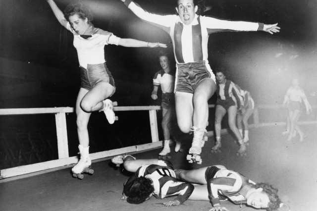 Black and white photograph of two women in roller skates jumping over two fallen skaters as they race around a wooden track.