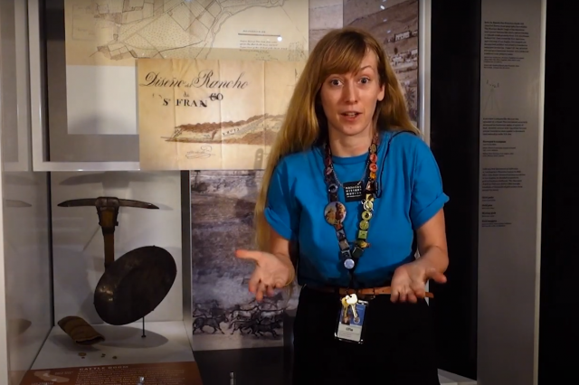 educator stands in front of exhibit case with gold pan and pick axe
