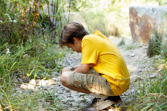 boy with light brown hair and a yellow shirt kneels and looks at something on the ground