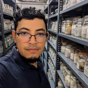 Scientist wearing glasses looks at camera amid jars of specimens on shelves